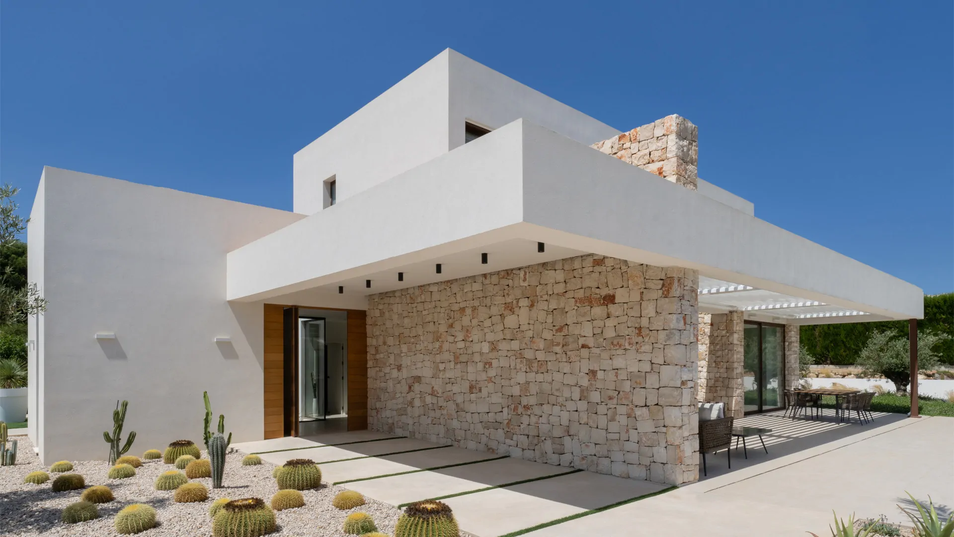 Una villa moderna de dos plantas con fachada blanca y una pared lateral de piedra texturizada. La villa cuenta con grandes ventanales y una pérgola que cubre una zona de comedor exterior.