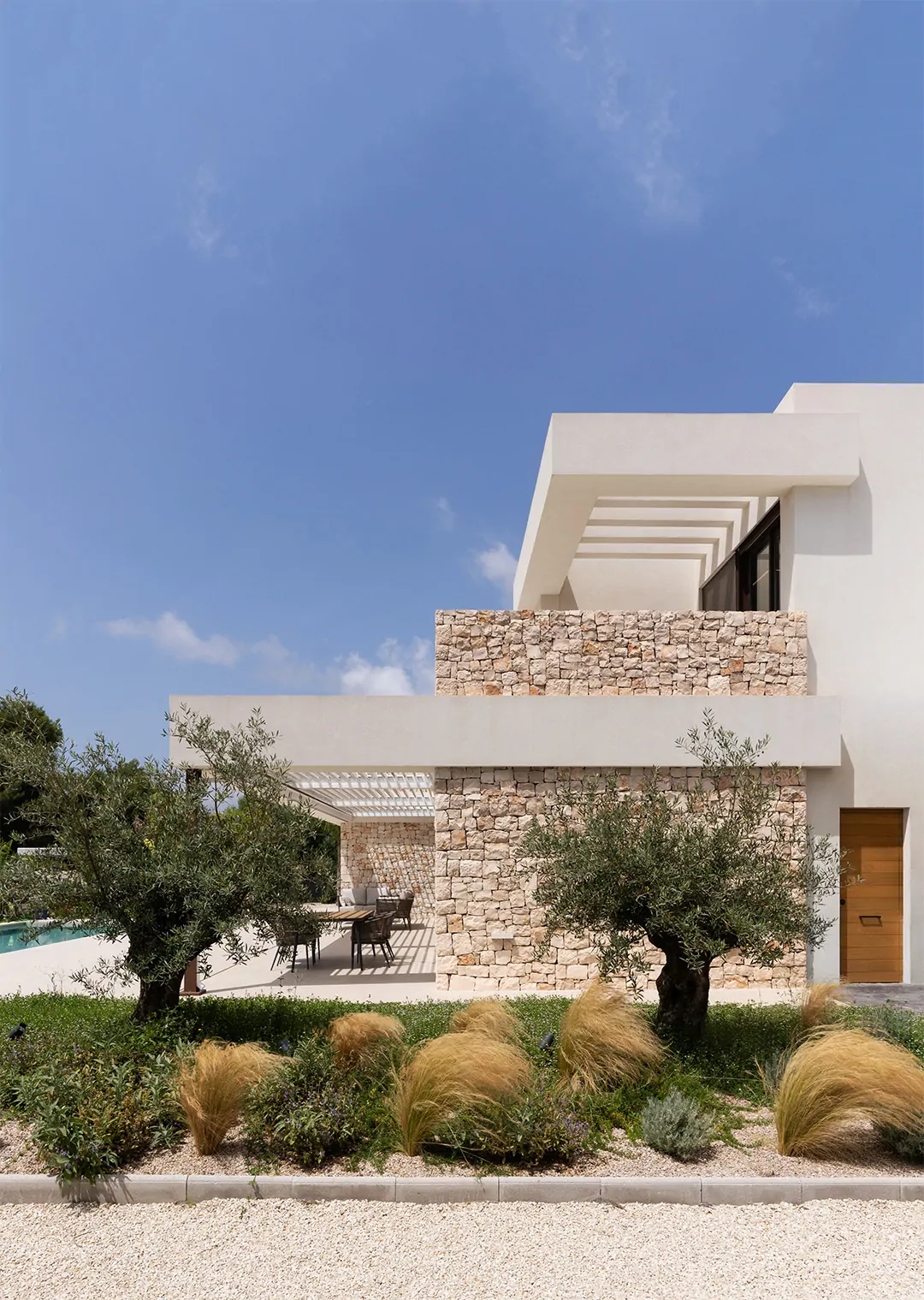 Una villa moderna de dos plantas con fachada blanca y una pared lateral de piedra. La villa cuenta con una pérgola que cubre una terraza en la planta superior y otra pérgola en la planta baja que cubre un comedor exterior.