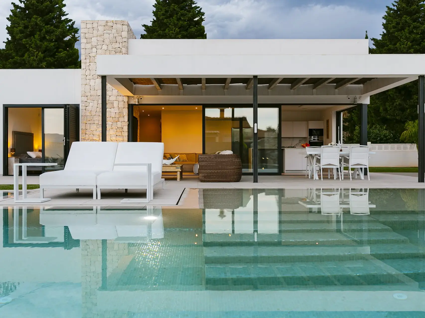 Una villa moderna de una planta con fachada blanca y detalles en piedra. La casa tiene grandes ventanales que dan a una terraza con piscina desbordante. En la terraza hay un sofá cama doble de color blanco y una mesa auxiliar del mismo color.