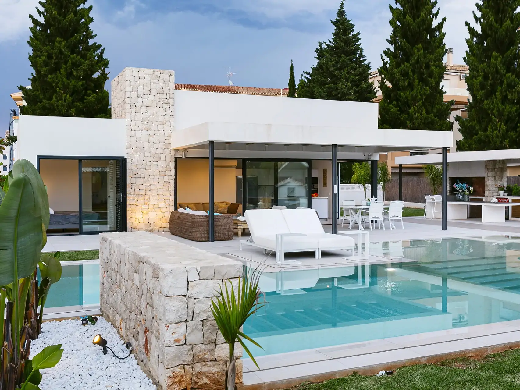 Una villa moderna de una planta con fachada blanca y detalles en piedra. La casa tiene grandes ventanales que se abren a una terraza con piscina desbordante. La terraza está amueblada con tumbonas, sofás y una mesa de comedor.