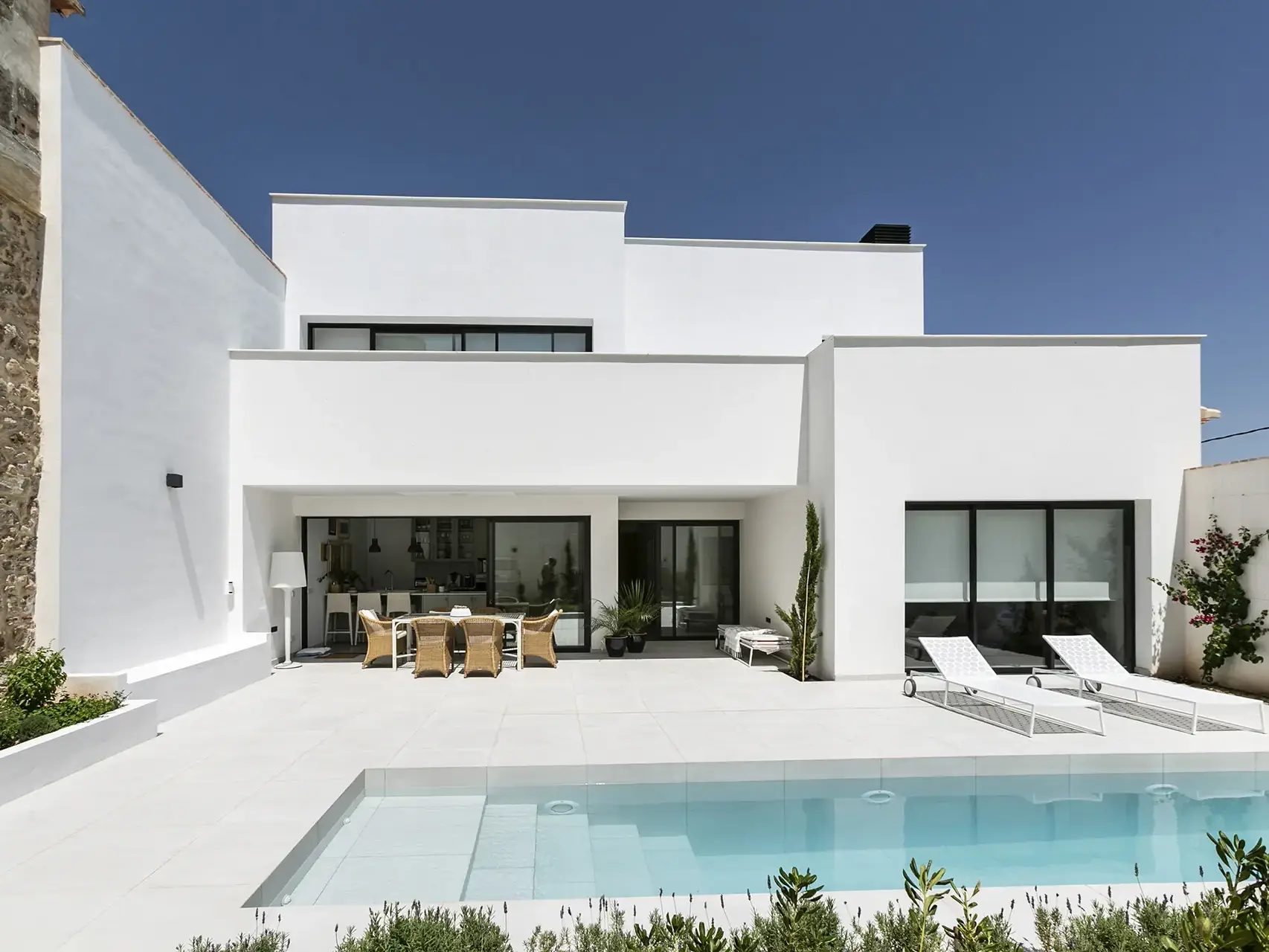 Una villa moderna de dos plantas con fachada blanca y detalles en piedra. La planta baja cuenta con grandes ventanales que se abren a un patio con piscina, tumbonas y un comedor exterior. El jardín está decorado con plantas y arbustos.