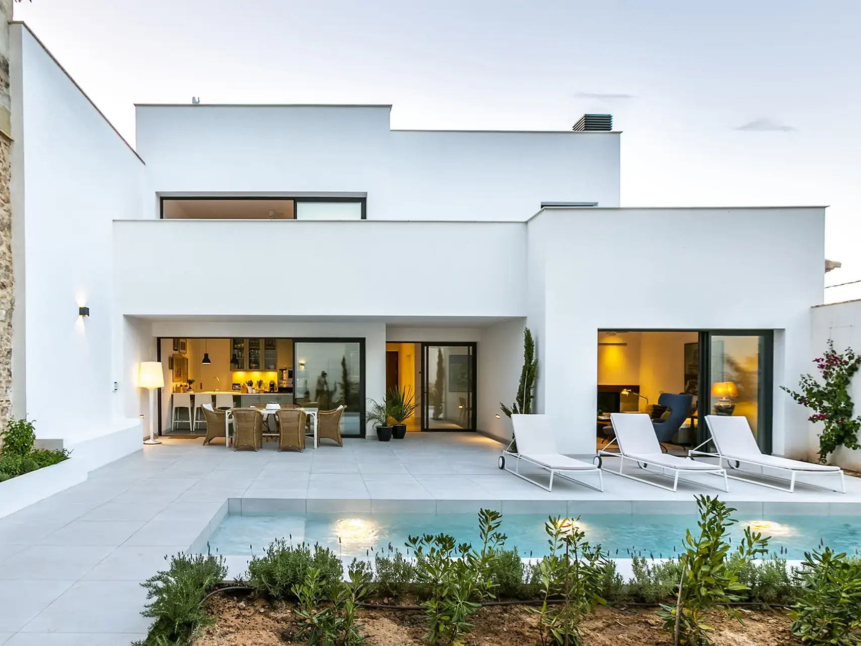 Una villa moderna de dos plantas con fachada blanca y una pared lateral de piedra. La planta baja cuenta con grandes ventanales que se abren a un patio con piscina, tumbonas y un comedor exterior. El jardín está decorado con plantas y arbustos.