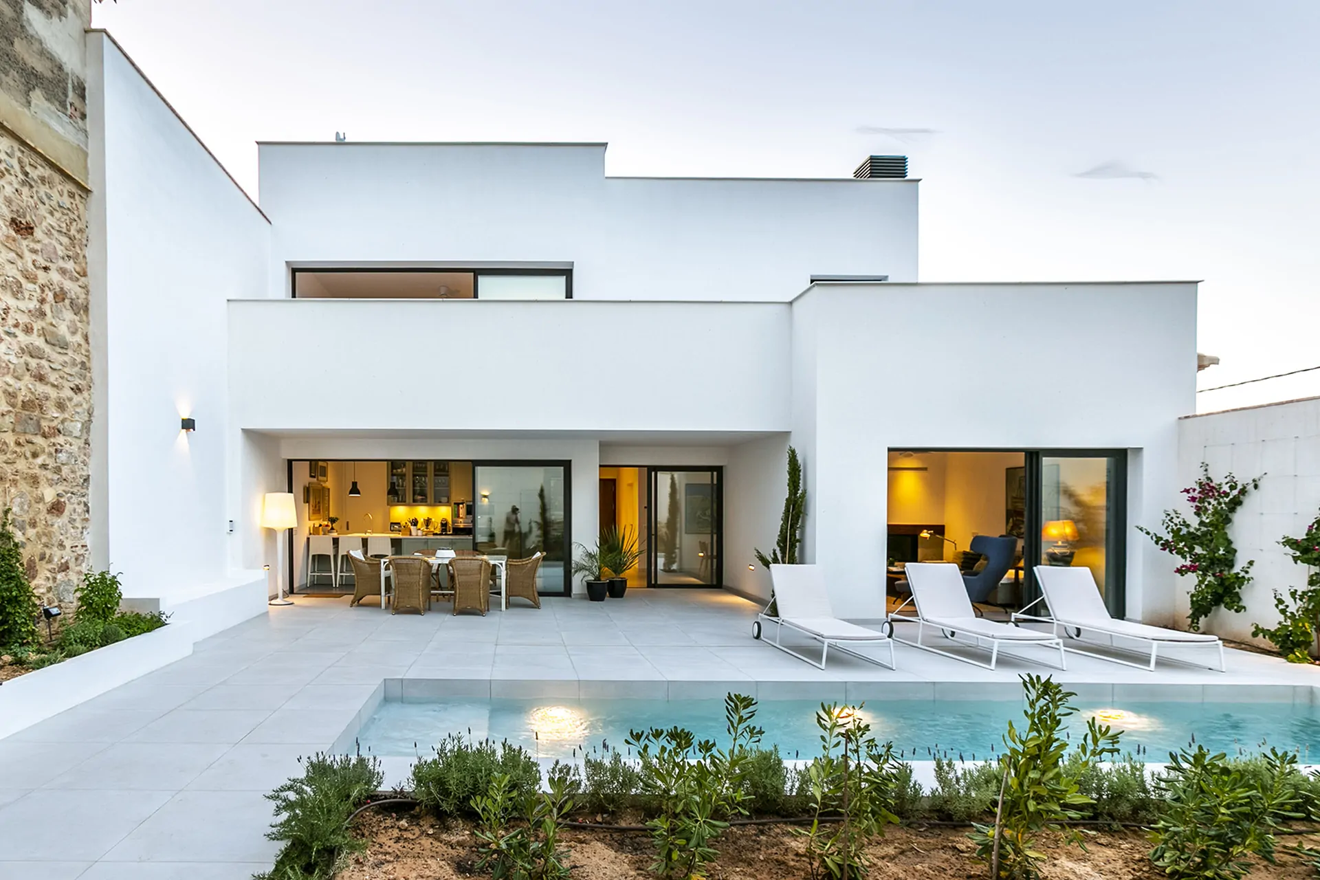 Una villa moderna de dos plantas con fachada blanca y una pared lateral de piedra. La planta baja cuenta con grandes ventanales que se abren a un patio con piscina, tumbonas y un comedor exterior. El jardín está decorado con plantas y arbustos.