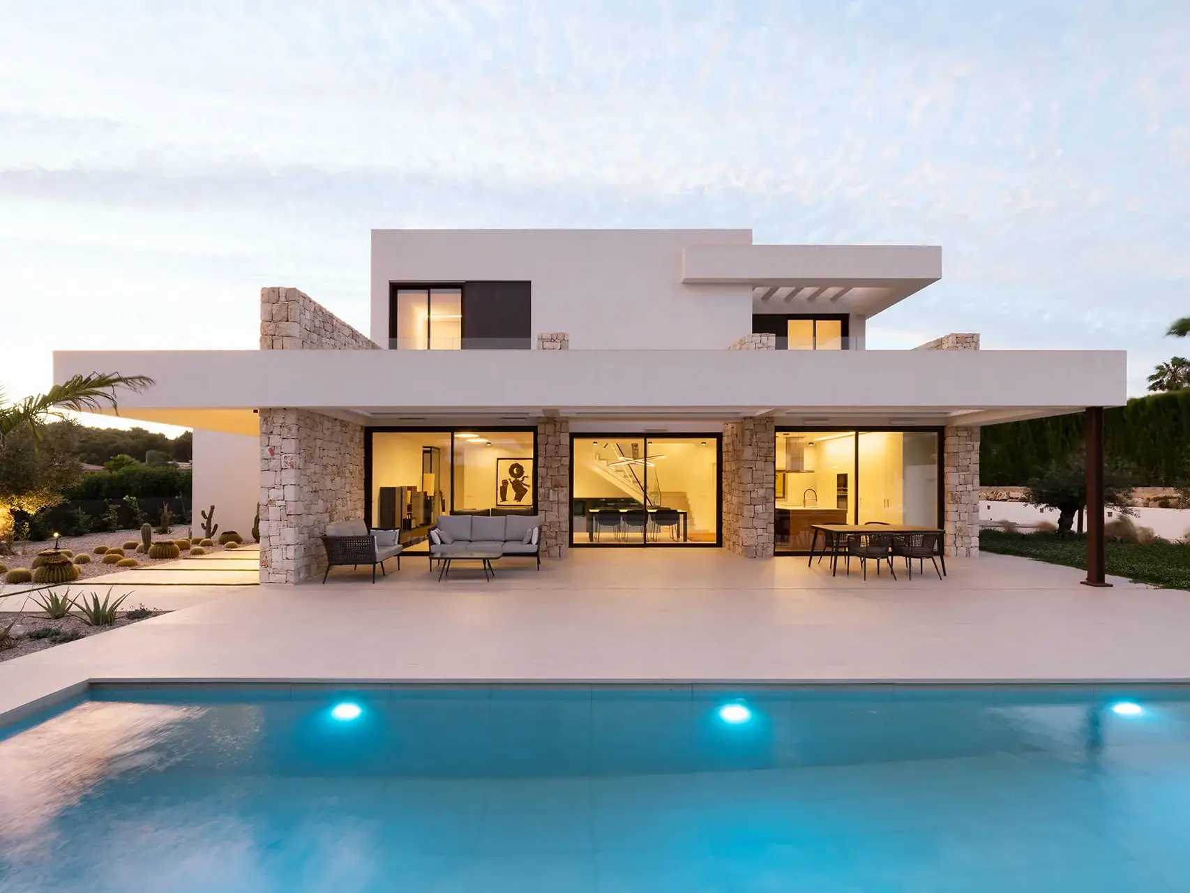 Una villa moderna de dos plantas con fachada blanca y detalles en piedra. La villa cuenta con grandes ventanales que se abren a una terraza cubierta.