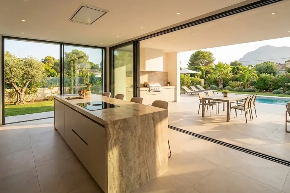 Ejemplo de cocinas abiertas al exterior mediante grandes ventanales correderos que conectan con la piscina en una villa de Jávea