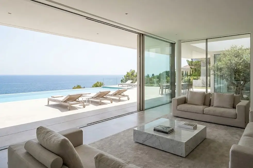 Salón de lujo con carpintería minimalista, grandes ventanales correderos de suelo a techo que conectan con el mar sin marcos visibles.